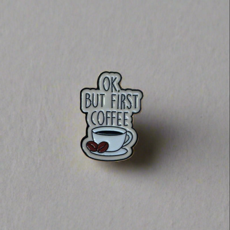 Hand holding a sticker with text 'OK, BUT FIRST COFFEE' against a blurred outdoor background.