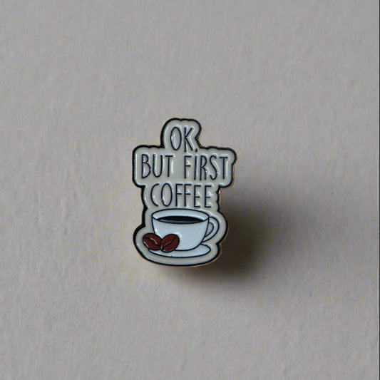 Hand holding a sticker with text 'OK, BUT FIRST COFFEE' against a blurred outdoor background.